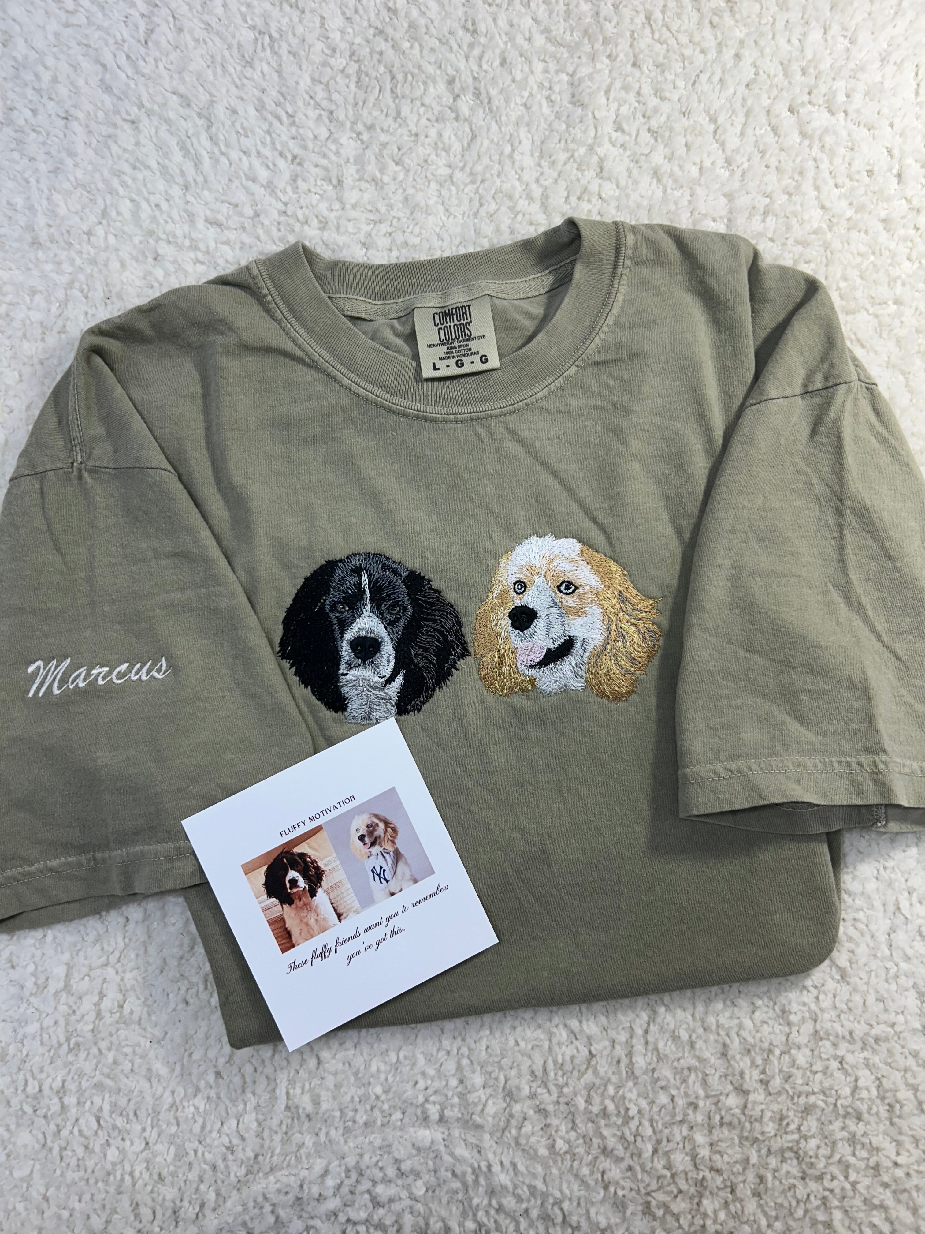 An Kaki short-sleeve t-shirt laid flat, featuring two embroidered dog faces on the front (one golden, one black and white) and a white card next to it.